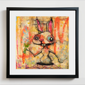 A framed surreal art print featuring a cartoonish rabbit holding a carrot, set against a colorful abstract background. The artwork is matted with white and enclosed in a black frame, shown hanging on a light gray wall.