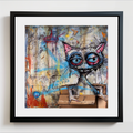 Framed wall art featuring a surreal painted cat with large red-and-blue eyes, pink ears, and a cardboard box the cat is in, set in a black frame with a white mat against a light wall.