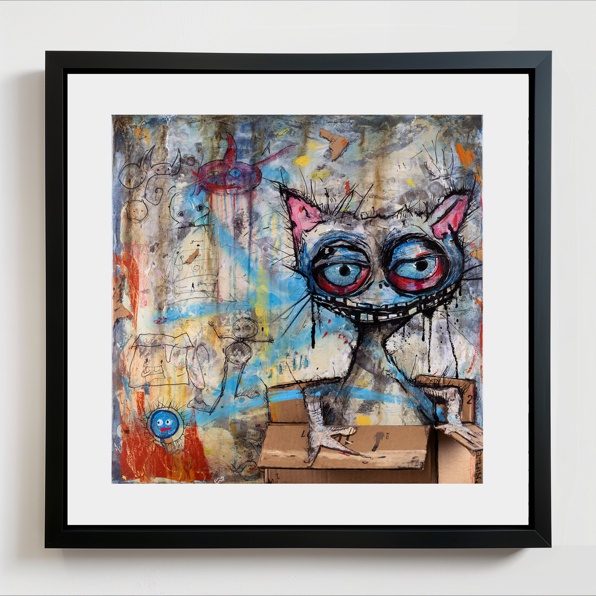 Framed wall art featuring a surreal painted cat with large red-and-blue eyes, pink ears, and a cardboard box the cat is in, set in a black frame with a white mat against a light wall.