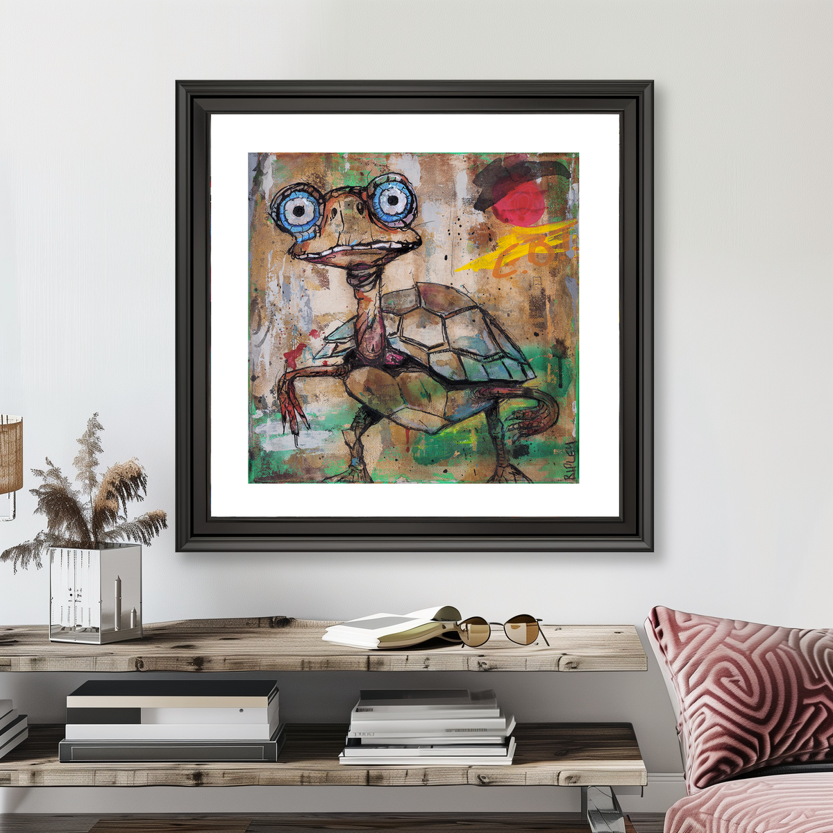 A framed abstract art print featuring a stylized turtle with large blue eyes hangs on a light gray wall above a wooden console table with books, sunglasses, and dried grass decor.