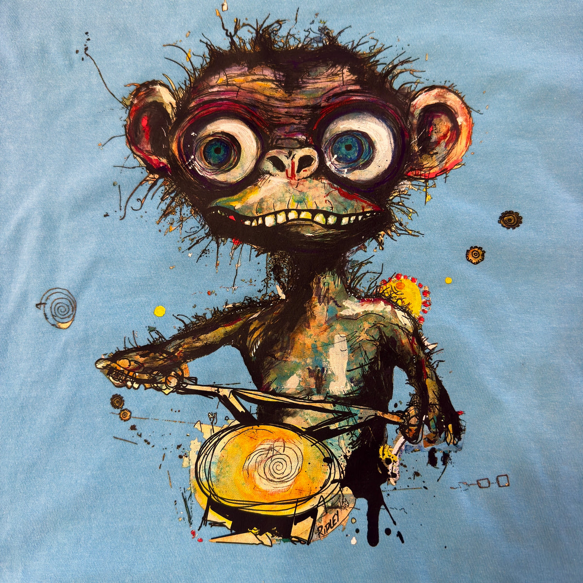 “Simian…ridin’ T-shirt graphic by Ripley, showing a wide-eyed monkey holding handlebars on bright blue fabric.” A close-up of a light blue fabric surface featuring a colorful, textured graphic print of a stylized monkey riding a bicycle.