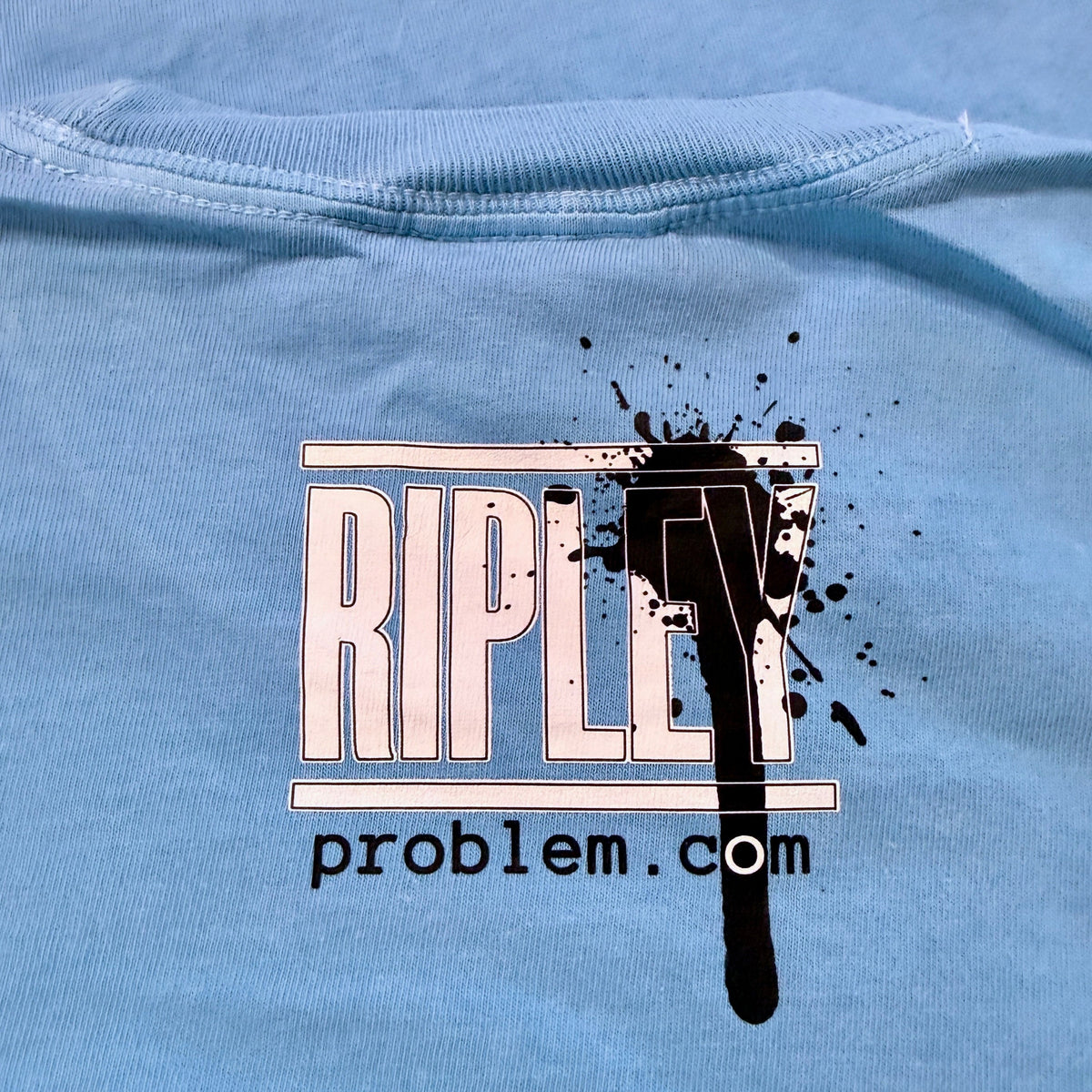 “Simian…ridin’ T-shirt graphic by Ripley, ”Close-up of a light blue t-shirt featuring a graphic print with the word 'RIPLEY' in white outlined letters, overlaid with a black paint splatter and drip, and the text 'problem.com' underneath. ripleyproblem.com