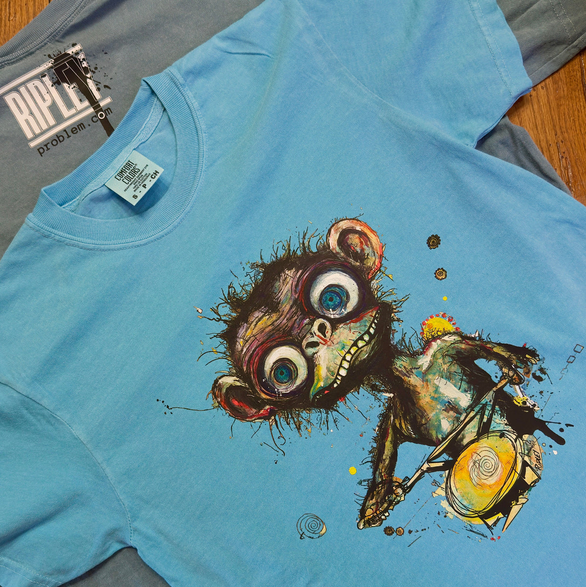 A light blue t-shirt featuring a detailed graphic print of a cartoon monkey with large blue eyes and a mischievous grin. The shirt is laid flat on a wooden surface, with a gray t-shirt underneath and a 'RIPLEYproblem.com.
