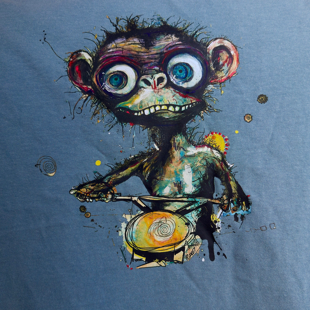 “Simian…ridin’ T-shirt graphic by Ripley, showing a wide-eyed monkey holding handlebars on dark blue fabric.”A close-up of a blue fabric garment featuring a colorful graphic print of a stylized, expressive cartoon monkey riding a bicycle.