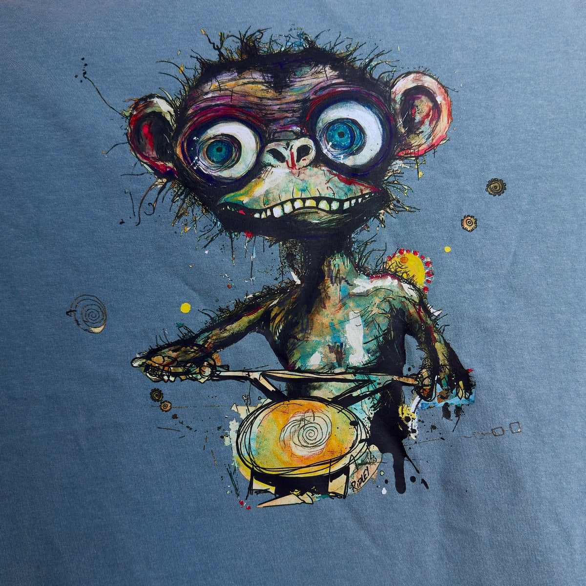 “Simian…ridin’ T-shirt graphic by Ripley, showing a wide-eyed monkey holding handlebars on dark blue fabric.”A close-up of a blue fabric garment featuring a colorful graphic print of a stylized, expressive cartoon monkey riding a bicycle.