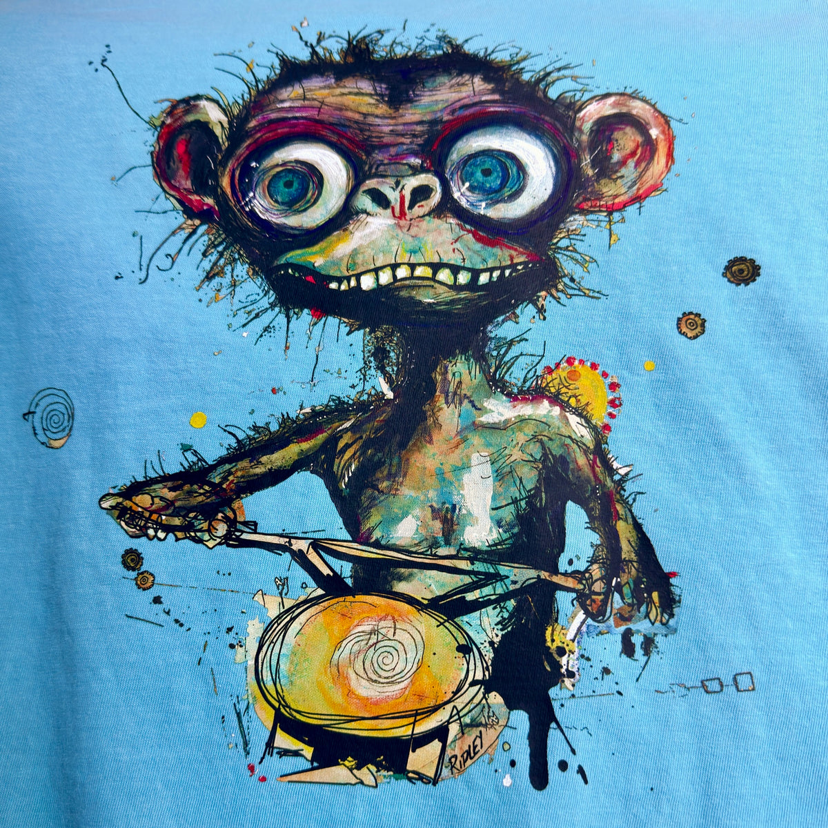 “Simian…ridin’ T-shirt graphic by Ripley, showing a wide-eyed monkey holding handlebars on bright blue fabric.” A light blue t-shirt featuring a detailed graphic print of a stylized, expressive cartoon monkey riding a dirt bike, with colorful paint splatters and expressive eyes.