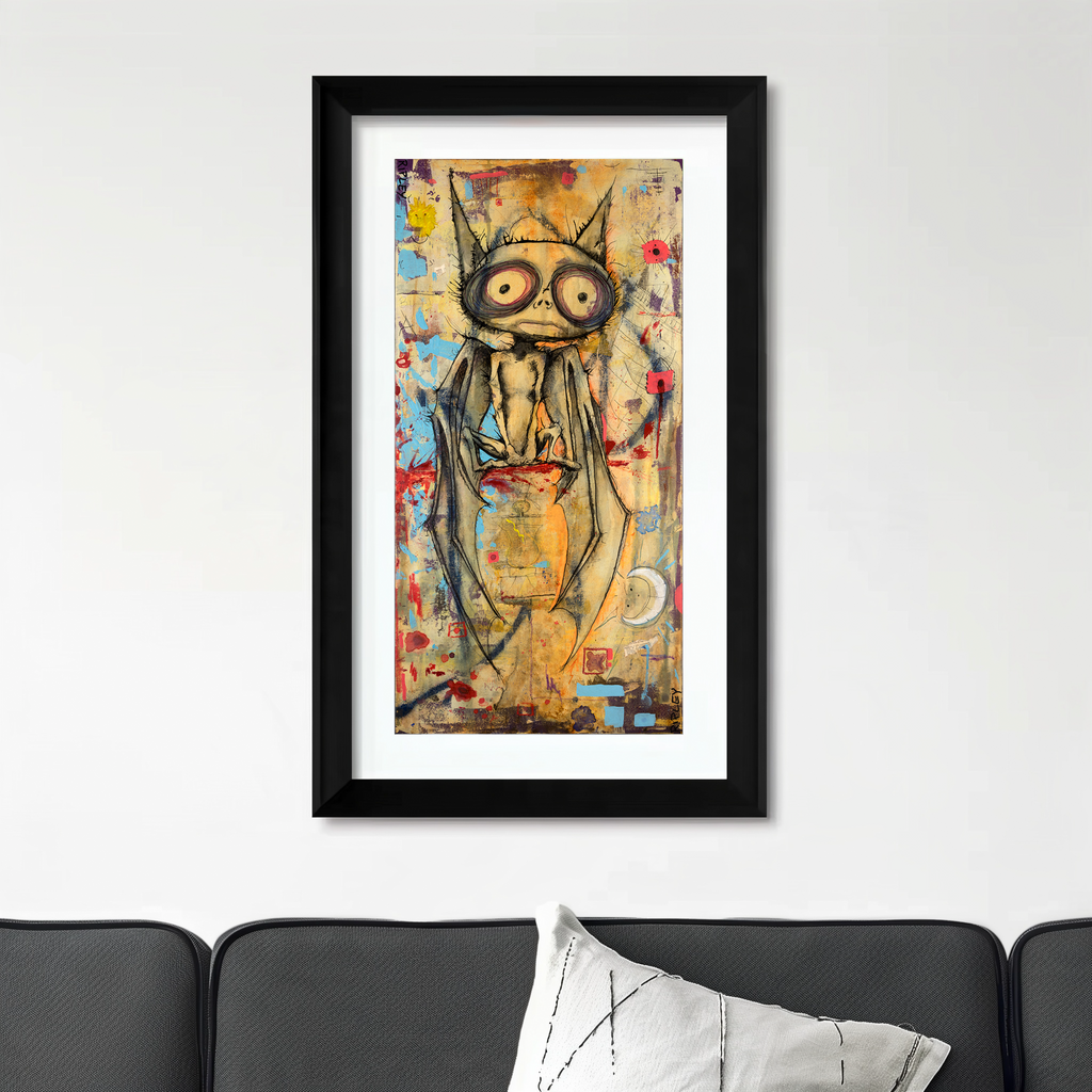 A framed vertical art print featuring a stylized bat illustration in black, gold, and red tones, displayed on a white wall above a dark gray sofa with a white pillow.
