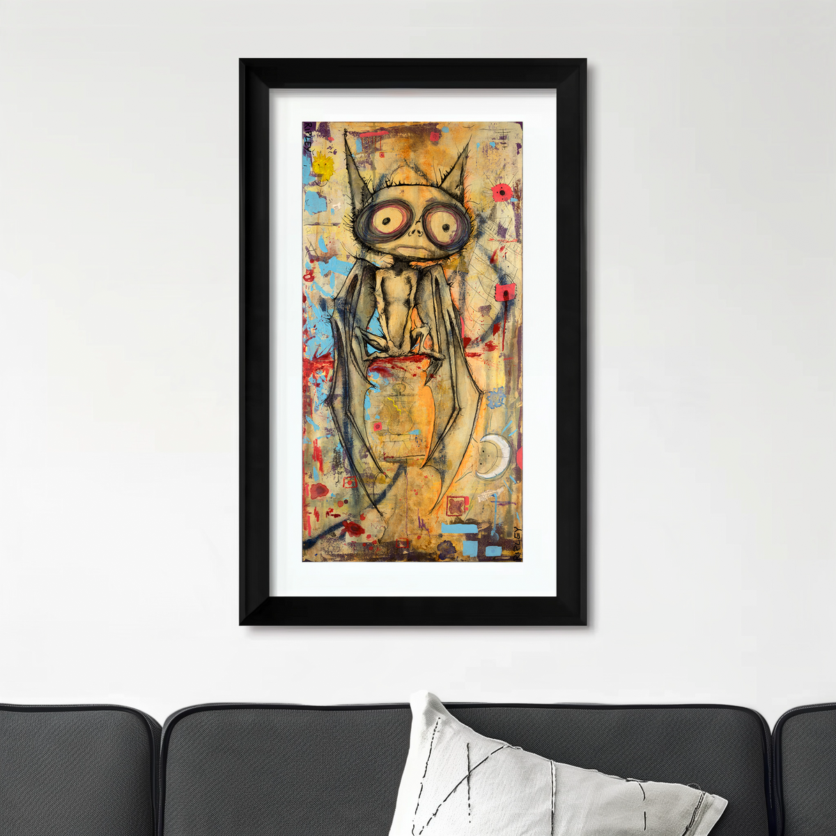 A framed vertical art print featuring a stylized bat illustration in black, gold, and red tones, displayed on a white wall above a dark gray sofa with a white pillow.