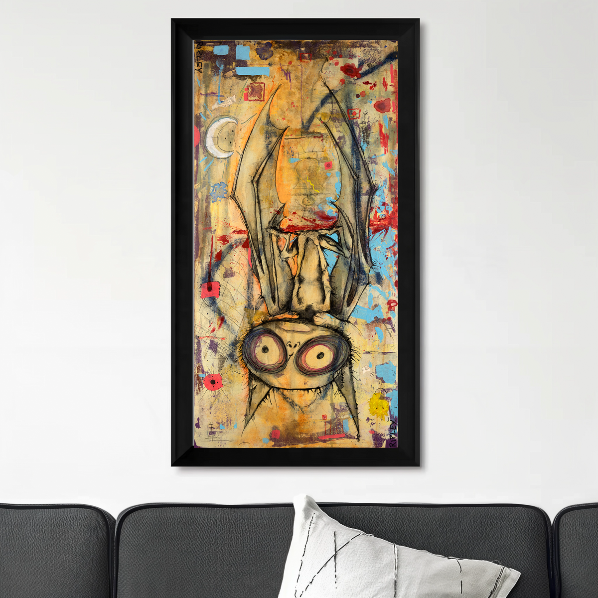 A framed abstract art print featuring a stylized bat figure with large eyes, hanging on a white wall above a dark gray sofa with a white pillow.