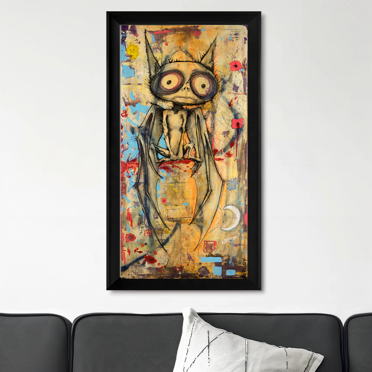 A framed abstract art print featuring a stylized bat with large eyes and bat-like wings, displayed on a white wall above a dark gray sofa with a white pillow.