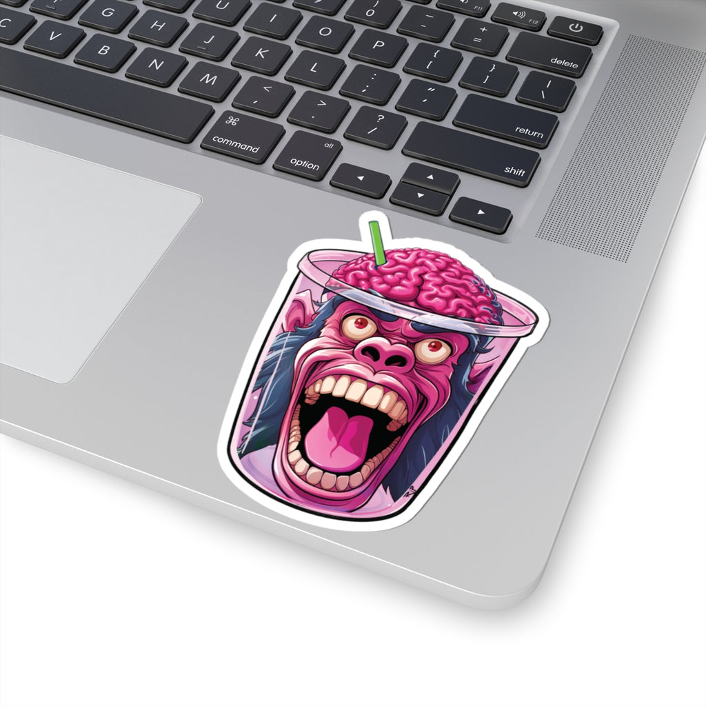 A die-cut vinyl sticker of a screaming pink gorilla with a brain for a hat inside a cup, shown applied to the silver palm rest of a laptop keyboard.