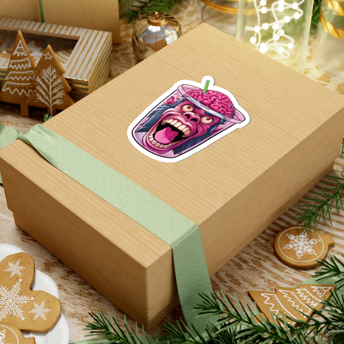 A kraft paper gift box tied with a pale green ribbon, featuring a colorful sticker of a screaming brain in a milkshake on top. The box is styled on a rustic wooden surface with Christmas cookies, pine branches, and holiday decorations.