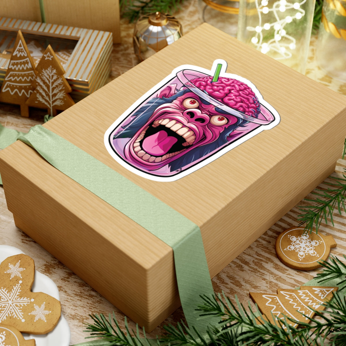 A kraft paper gift box wrapped with a pale green ribbon, decorated with a colorful sticker of a screaming pink brain character in a drink cup. The box is styled on a rustic wooden table with Christmas cookies, pine branches, and holiday decorations.