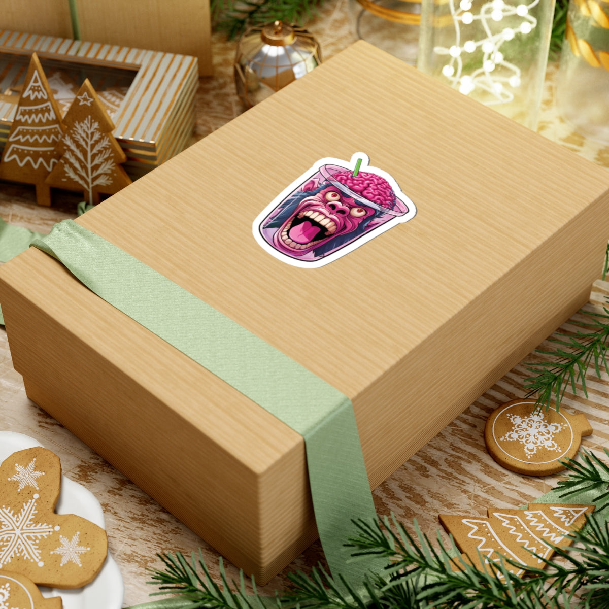 A kraft paper gift box tied with a pale green ribbon, featuring a colorful sticker of a pink monster face on top, styled on a wooden surface with holiday decorations.