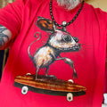 A close-up of a person wearing a bright red t-shirt with a graphic print of a creepy, large-eyed mouse balancing on a skateboard. The person also wears a black beaded necklace.