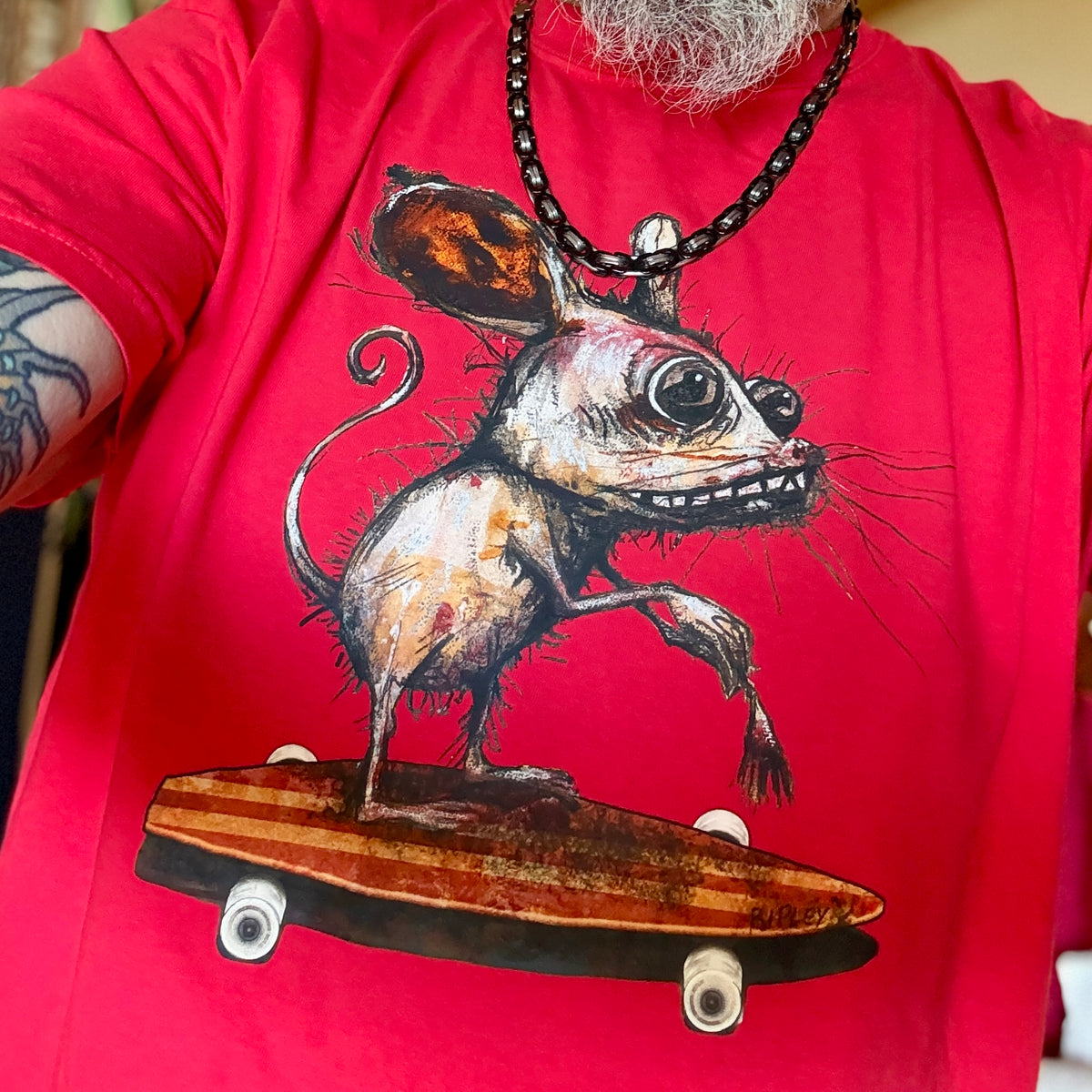 A close-up of a person wearing a bright red t-shirt with a graphic print of a creepy, large-eyed mouse balancing on a skateboard. The person also wears a black beaded necklace.