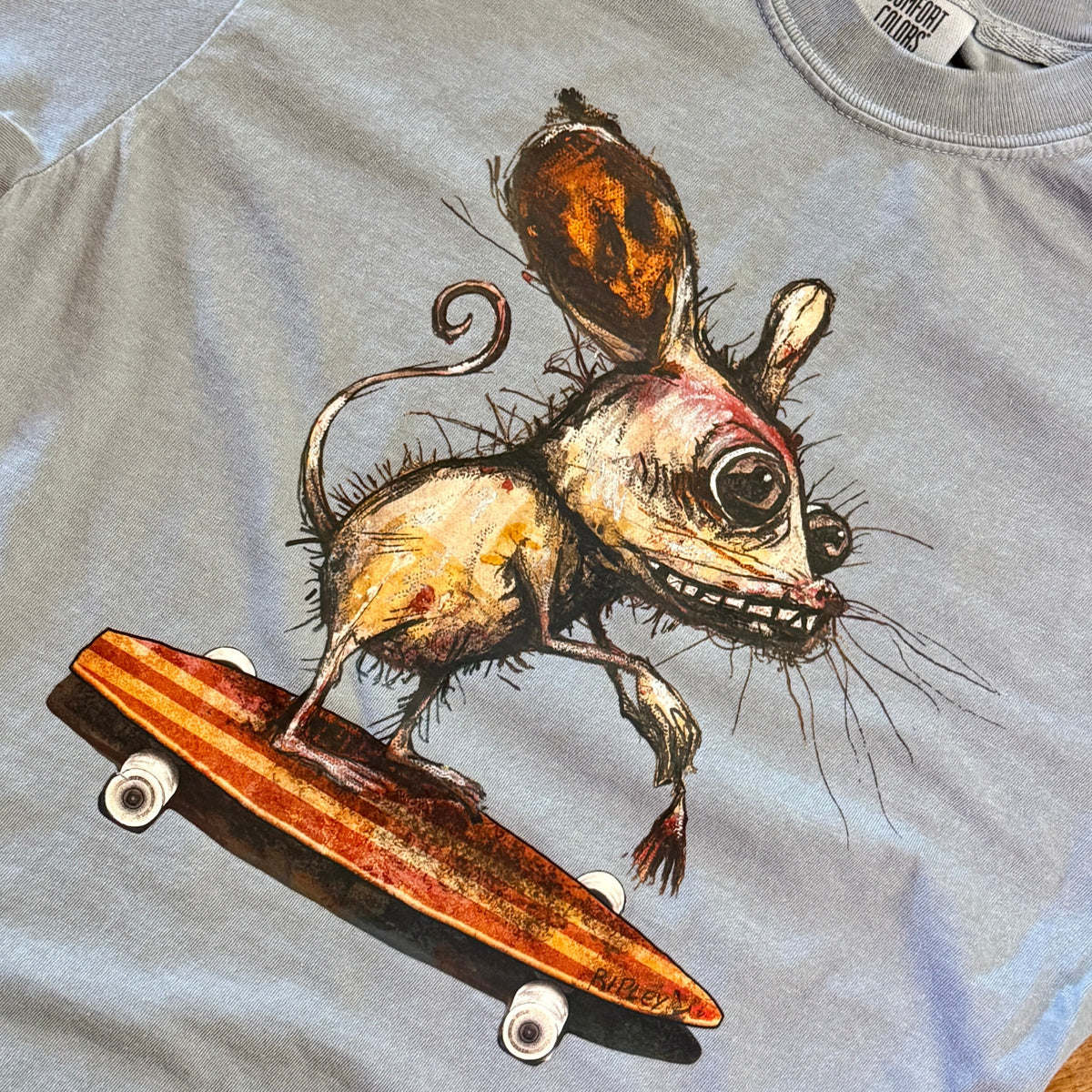 A close-up of a light gray t-shirt featuring a detailed graphic print of a creepy, stylized mouse riding a skateboard. The shirt fabric and collar are visible, with a brand tag partially visible at the top right.