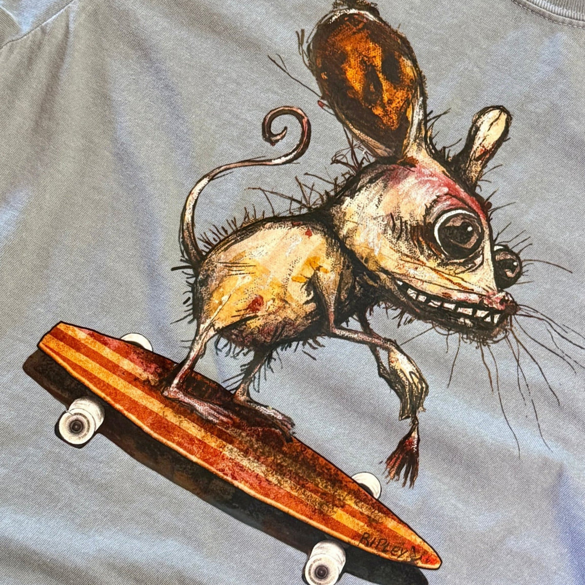 A grey crewneck t-shirt featuring a detailed graphic print of a creepy, stylized mouse riding an orange skateboard, laid flat on a wooden surface.