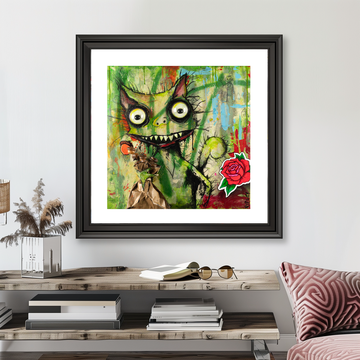 A framed abstract art print featuring a stylized green squirrel  face with large eyes and a red rose, displayed on a light gray wall above a wooden console table with books and decor.