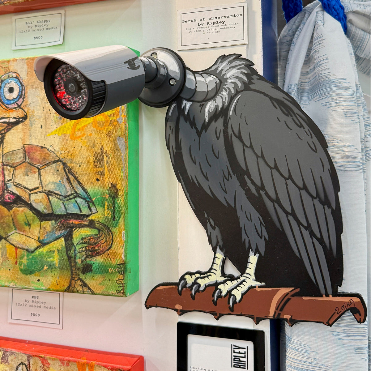 A large wall-mounted sculpture of a dark bird of prey perched on a branch, with a security camera mounted on its head, displayed against a white wall with colorful framed artwork nearby.