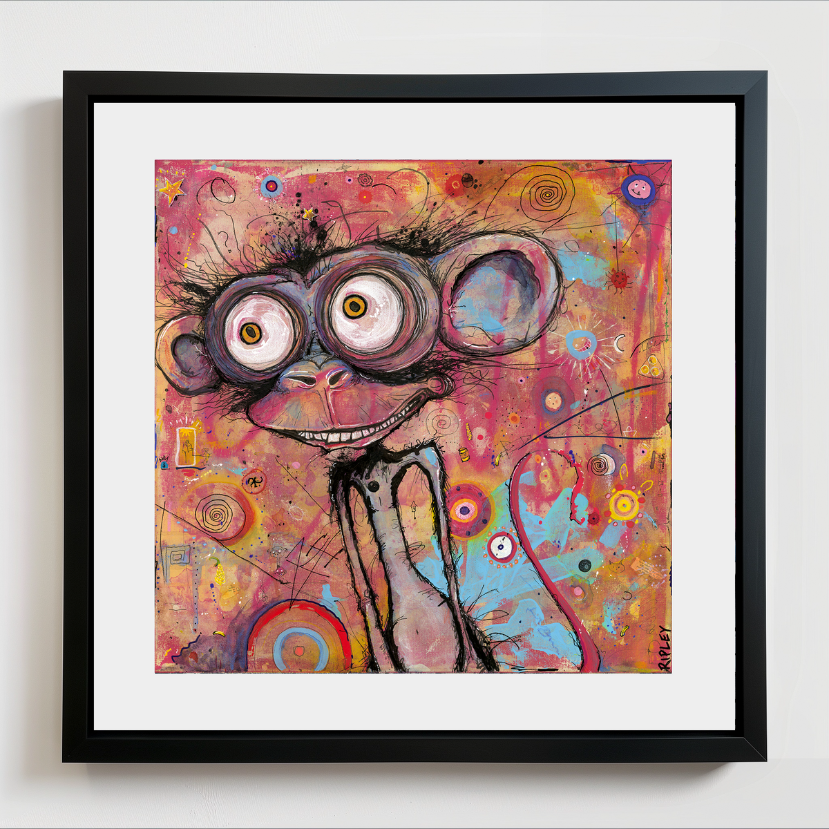 A framed abstract art print featuring a whimsical, cartoonish monkey face with oversized eyes and a wide grin, rendered in a colorful mix of pink, orange, yellow, blue, and purple tones. The artwork is presented in a black frame with a white mat against a clean white wall.